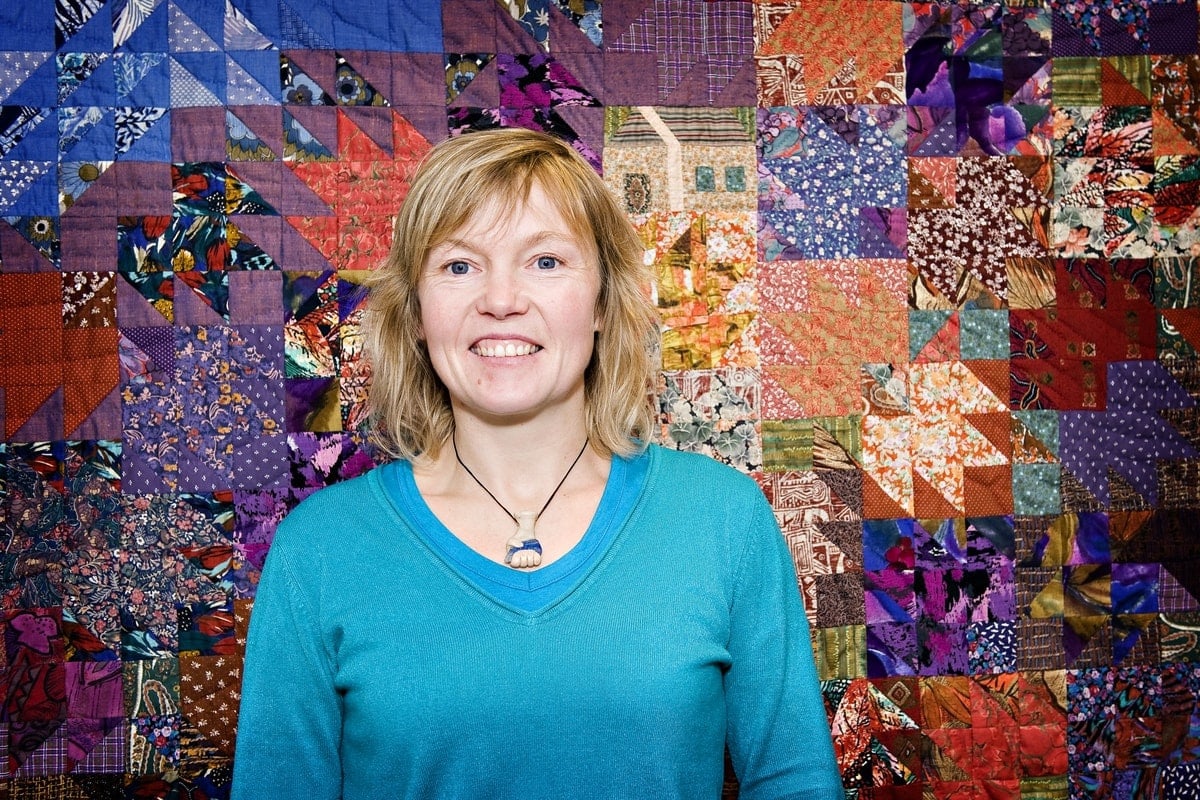 Open Aye. a portrait of a woman who stands in front of a colourful tapestry on the wall behind her.