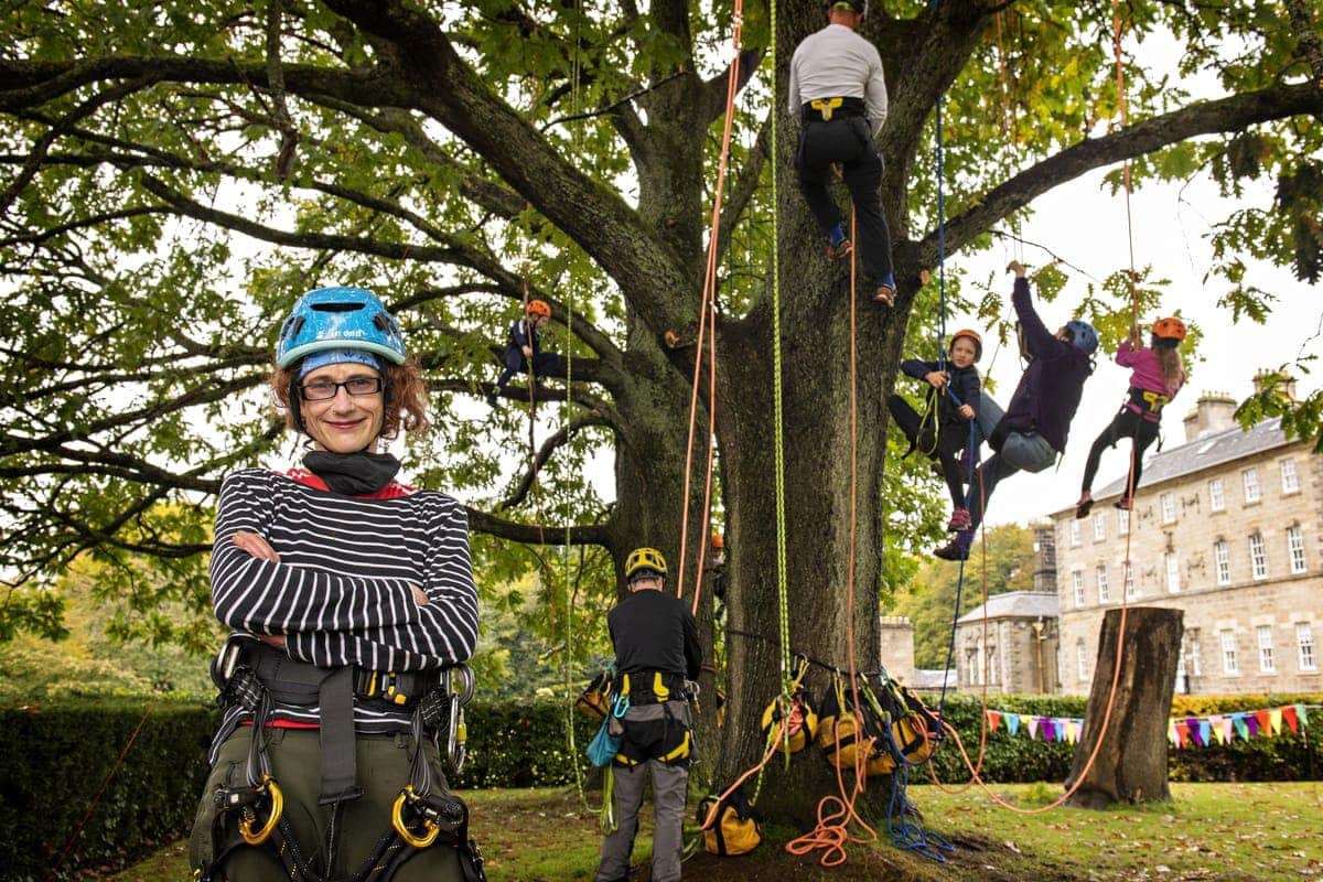 Open Aye. a woman poses for the camera whilst a group of children wearing helmets and harnesses climb a tree behind her.