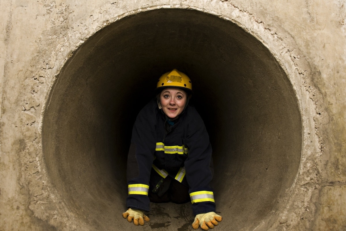Open Aye. a woman climbs through a tunnel wearing a helmet, gloves and a boiler suit.