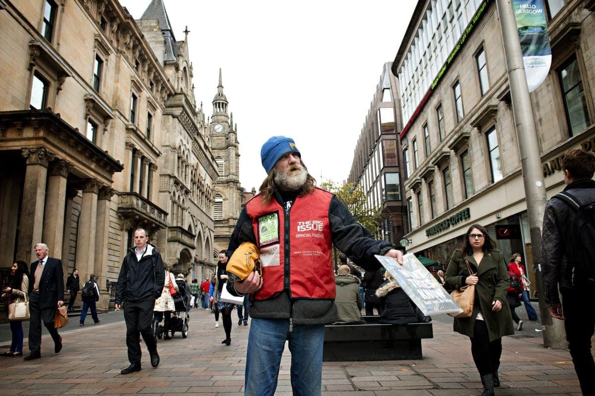 Open Aye. a man hands out the big issue magazine on a busy street in a city.