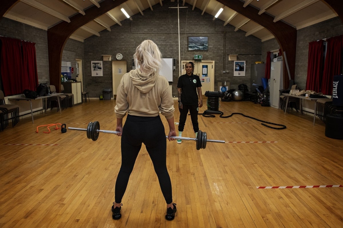 Open Aye. a woman lifts a barbell in a hall with wooden floors.