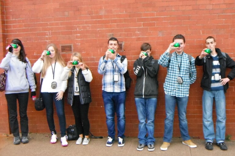 Open Aye. A group of people holding a camera up to their faces whilst standing against a brick wall.