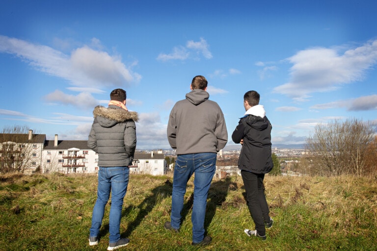 Open Aye. Photo of three men standing in a field looking onto a cityscape.